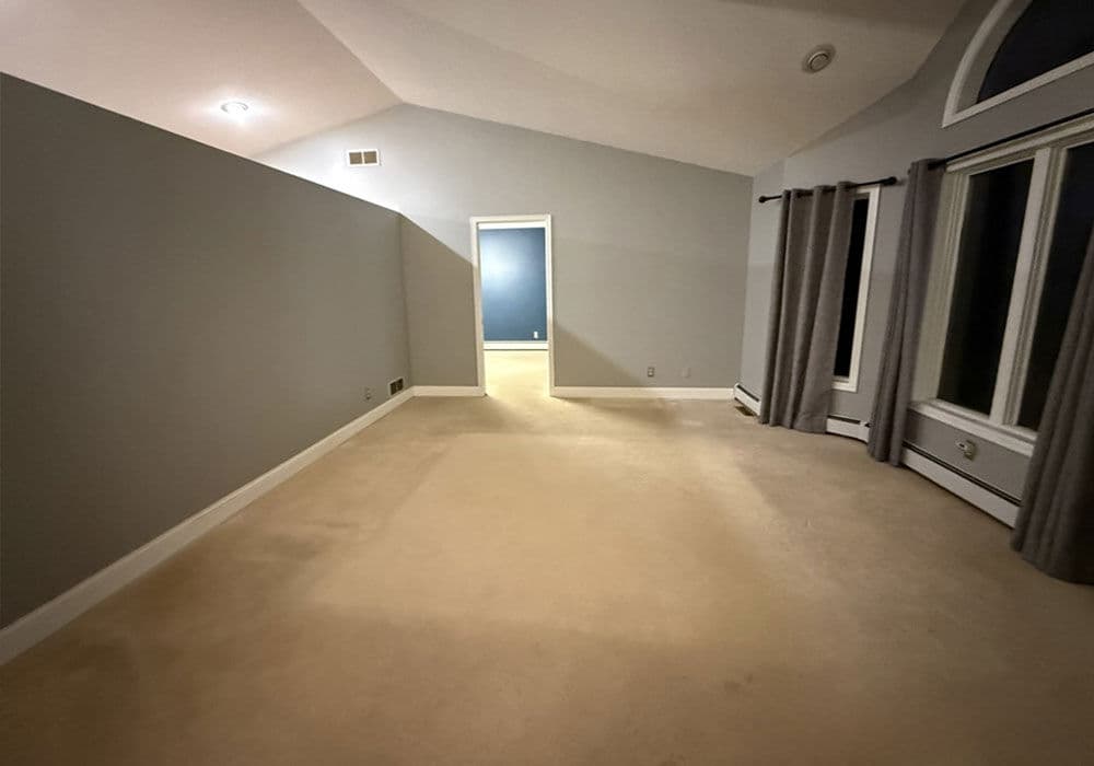 Spacious empty living room with beige carpet and gray walls, featuring large windows and vaulted ceiling.