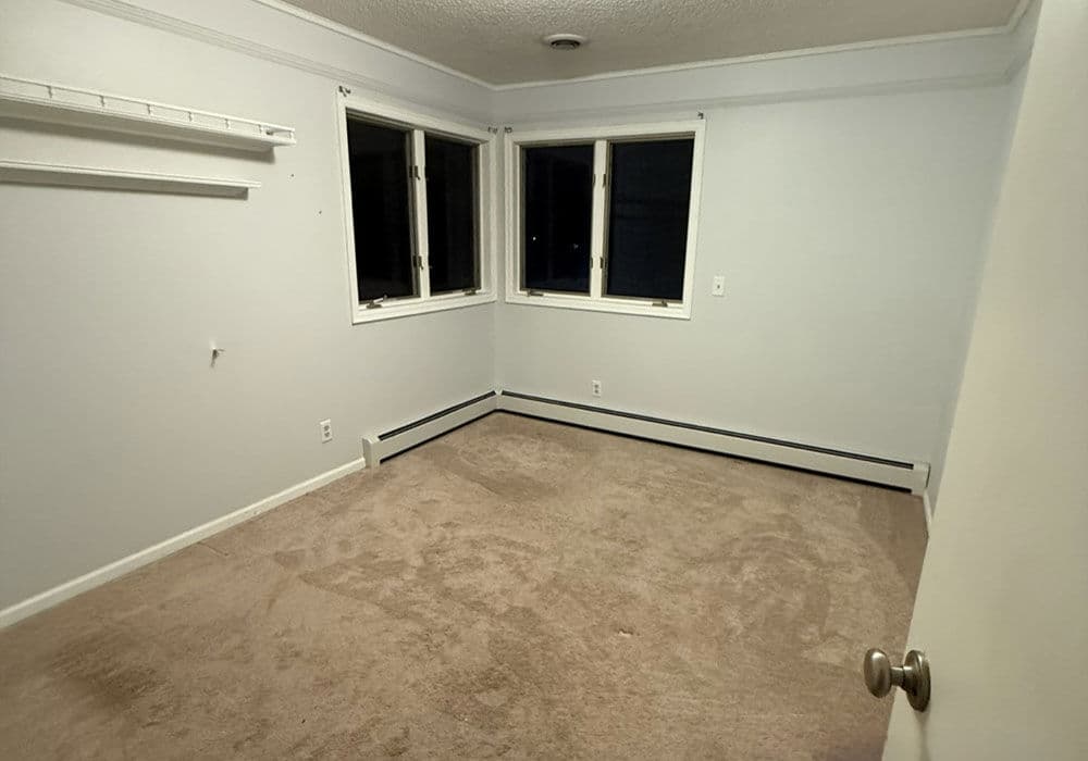 Empty beige room with carpet, white walls, and two windows, ideal for potential home buyers.